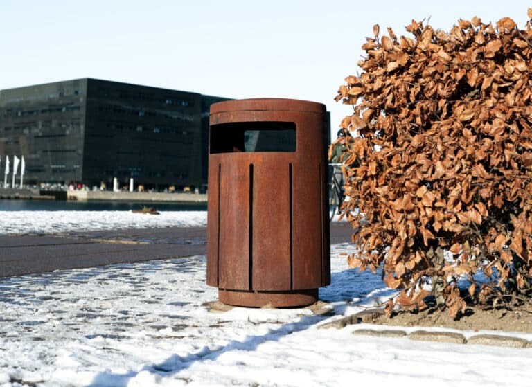 Rusten affaldsbeholder ved busk i sne.